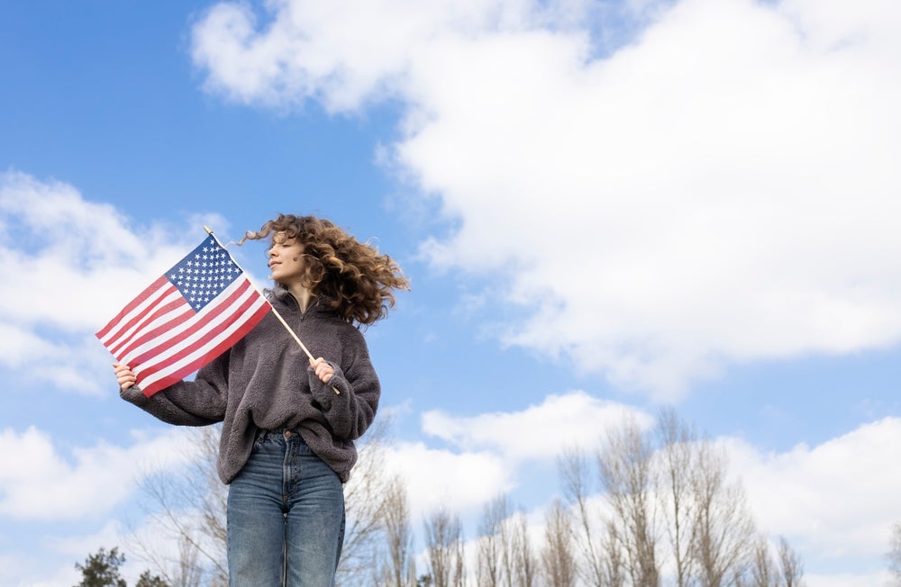 A young girl holding an American flag with a cloudy blue sky overhead