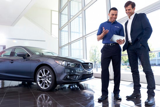 two man stand together looking at a pad of paper in front of a gray mazda 