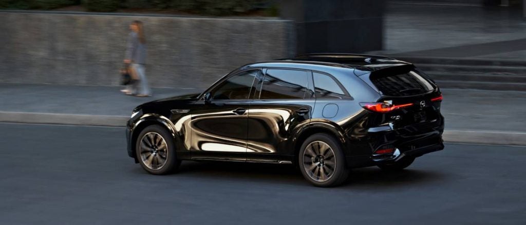 a black mazda suv driving on a road while a woman walks on the sidewalk beside it 