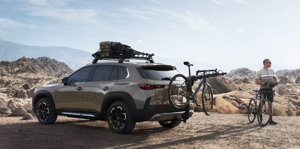a taupe suv is parked on a rocky landscape the biking equipment strapped to it and a man stands next to the car with a bike