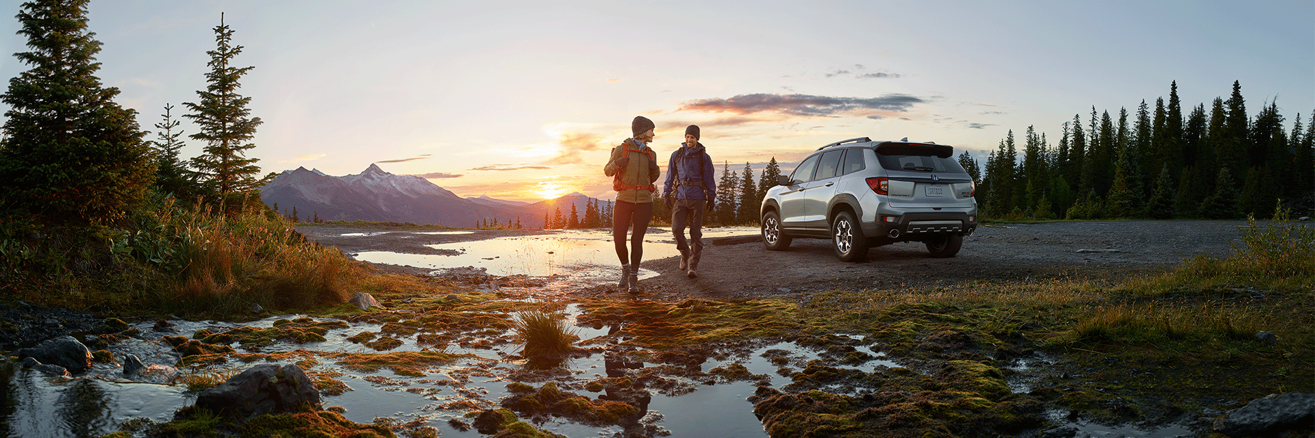Bennett Honda of Lebanon is a Honda Dealership in Lebanon near Palmyra, PA | People in Hiking Gear Walking away from 2022 Silver Honda Passport towards Camera with Sun Setting Behind Mountains and Lake in Background