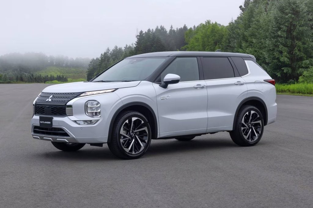 Silver Mitsubishi Outlander SUV with a black roof and black wheels on asphalt, trees and fog in the background.