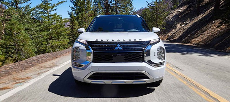 Front view of a white 2022 Mitsubishi Outlander SUV driving on a paved road through a forest.