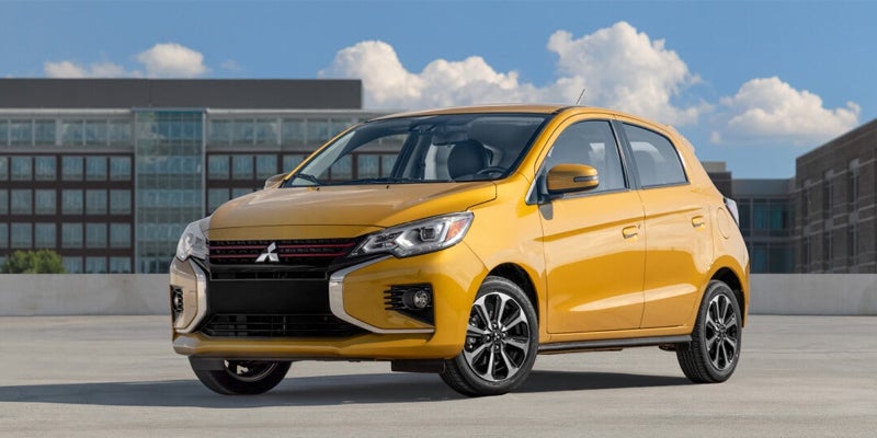 A gold Mitsubishi Mirage hatchback parked outdoors, angled toward the left, with buildings and blue sky with clouds in the background.