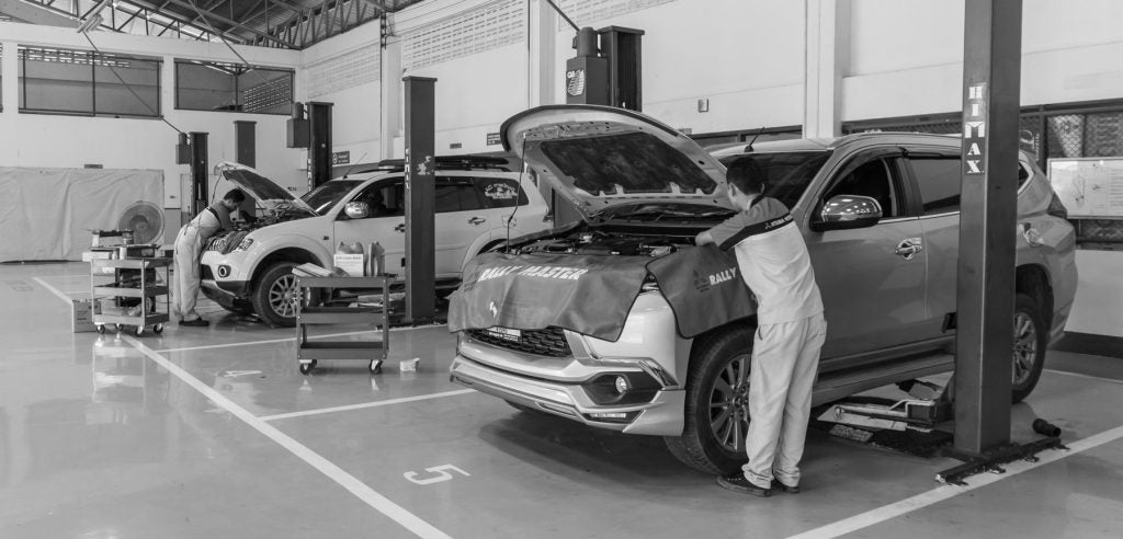 Two mechanics work on cars with open hoods in a brightly lit garage with car lifts.