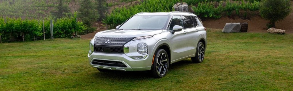 Silver Mitsubishi Outlander SUV parked on a green lawn with a vineyard and trees in the background.