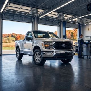 Professional automotive photography, low-angle heroic shot of a pristine metallic silver 2026 Ford F-150 pickup truck inside a state-of-the-art, sun-drenched dealership service bay, the truck is positioned on a polished charcoal concrete floor, cinematic overhead LED lighting reflecting off the clean body panels and chrome accents, a technician in professional workwear is visible in the background near a diagnostic computer, through large glass bay doors the gently rolling hills of a rural Ohio landscape are visible under a clear autumn sky with vibrant orange and red foliage, atmosphere of reliability and precision, high-resolution, photorealistic, sharp focus on the truck’s front fascia and rugged tires, clean and modern aesthetic, cool blue and warm amber color palette.