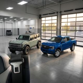 A high-detail, cinematic wide shot of a 2026 Ford Bronco and a 2026 Ford Ranger parked side-by-side inside a pristine, modern automotive service bay, polished concrete floors reflecting the bright overhead LED lights, the driver-side door of the Bronco is slightly ajar to reveal a premium interior with a focus on the silver seat belt buckle and black safety webbing, soft afternoon sunlight streaming through large glass garage doors in the background, a professional and reassuring atmosphere, vibrant Cactus Gray paint on the Bronco and Velocity Blue on the Ranger, sharp focus on the vehicle textures, realistic automotive photography, 8k resolution, clean and organized workshop environment, neutral color palette with pops of Ford blue.