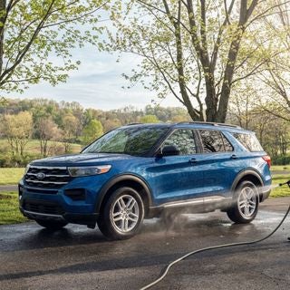 A pristine 2026 Ford Explorer in a vibrant, high-gloss finish, parked on a paved road in a quiet Calcutta, Ohio, late spring afternoon, soft May sunlight filtering through budding maple trees, a gentle mist from a garden hose rinsing the undercarriage, clear water droplets beading on the deep blue metallic paint, gently rolling hills damp asphalt, light spring clouds in a pale blue sky, crisp and clean atmosphere, eye-level perspective, cinematic lighting, sharp focus on the vehicle's rocker panels and wheel wells, cool and refreshing color palette. no houses in background