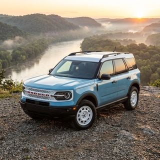 A 2026 Ford Bronco Sport Heritage in a vintage-inspired Robin’s Egg Blue with a signature Oxford White roof, parked on a scenic gravel overlook perched above the winding Ohio River, panoramic views of the Tri-State riverfront bluffs at dawn, soft morning mist rising from the water, lush deciduous forests and rolling Appalachian hills in the background, cinematic low-angle three-quarter shot, focus on the retro-style white wheels and classic grille, warm golden hour sunlight reflecting off the polished metallic paint, crisp textures of the off-road tires and rocky terrain, adventurous and nostalgic mood, photorealistic automotive photography, high-detail finish, no text or logos.