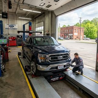 A wide-angle, eye-level shot of a clean 2023 Ford F-150 parked on a precision alignment rack inside a well-lit, professional service bay in East Liverpool, Ohio. The truck, in a practical Antimatter Blue, is the focal point, its front wheels perfectly straight as a technician in a grey Ford service uniform adjusts a sensor. Through the open garage bay doors, the bright, hazy sunlight of a warm May afternoon illuminates a realistic, modest commercial landscape typical of the Ohio River valley, featuring a glimpses of a paved lot, a weathered brick building across the street, and the lush, bright green foliage of late spring. The lighting is natural and airy, reflecting the transition to summer, with soft reflections on the truck's polished hood. The color palette is grounded in mechanical greys, tool-chest reds, and the deep blue of the truck, creating a mood of expert care and seasonal readiness for a hard-working community.