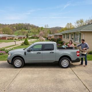 2026 Ford Maverick XL hybrid pickup truck, stone gray paint, parked on a paved driveway of a modest mid-century ranch-style home in Calcutta, Ohio, late spring morning, May 12th, bright diffused sunlight, clear blue sky with light wispy clouds, a family loading a tackle box and fishing rods into the truck bed, background featuring budding deciduous trees and rolling green hills typical of the Appalachian Plateau, a quiet suburban street with similar well-kept modest houses, clean and realistic lighting, reflections on the truck's bodywork, low-angle side-profile shot emphasizing the vehicle as the centerpiece, spring vegetation with small flowers blooming in a simple garden bed, authentic working-class suburban atmosphere, high detail, photographic style.