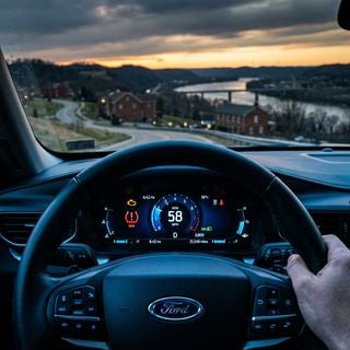 A cinematic, close-up driver’s perspective of a modern Ford digital instrument cluster, high-definition glowing warning icons including an engine silhouette and a tire pressure symbol illuminated in vibrant amber and red against a sleek black screen, shallow depth of field focusing on the crisp digital graphics, soft-focus background showing a steering wheel and a glimpse through the windshield of a hilly tri-state landscape with brick architecture and the Ohio River valley at dusk, cool blue interior ambient lighting contrasting with the warm golden hour light outside, photorealistic style, sharp textures, professional automotive photography, high-contrast, moody and informative atmosphere.