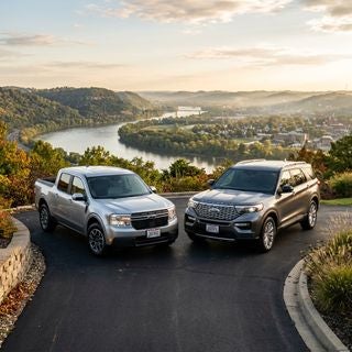 Cinematic automotive photography, a sleek 2026 Ford Maverick compact pickup and a sophisticated Ford Explorer SUV parked side-by-side on a clean asphalt driveway, dramatic low-angle perspective, overlooking the lush riverfront bluffs and rolling hills of the Ohio River valley during golden hour, warm sunlight glinting off the polished metallic paint and chrome accents, professional maintenance aesthetic, clean tires, pristine showroom condition, misty distant valley background with hints of regional architecture, vibrant greens and soft autumnal earth tones, soft lens flare, high dynamic range, crisp details on the intricate grilles and LED headlights, serene and dependable atmosphere, hyper-realistic textures, 8k resolution.