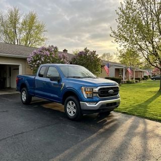 A rugged, metallic blue Ford F-150 parked in a clean, asphalt driveway of a modest, mid-century ranch-style home in Calcutta, Ohio, late afternoon sun casting long shadows across a well-kept green lawn, American flags on several neighboring porches for Memorial Day, flowering lilac bushes and budding maple trees in the background, soft spring light, overcast sky with hints of golden hour, wide-angle shot, ground-level perspective emphasizing the truck's chrome grille and muscular stance, realistic suburban street with no luxury upgrades, working-class neighborhood atmosphere, damp pavement reflecting the sky from a recent light spring shower, peaceful and patriotic mood, cinematic realism.