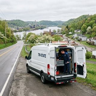 A wide-angle, eye-level shot of a clean, white Ford service van that says tristate on the side parked on the asphalt shoulder of a quiet, two-lane road in East Liverpool, Ohio, on a mild, overcast Friday in early May. In the background, the industrial silhouettes of the Ohio River waterfront and modest, mid-20th-century working-class homes sit nestled among gently rolling hills with fresh spring greenery and blooming hawthorn trees. A professional technician in a dark blue Ford service uniform is neatly organizing tools inside the rear of the van, the focus on the efficiency of the mobile service. The lighting is soft and diffused from the grey spring sky, creating realistic reflections on the van's polished white paint. The overall mood is dependable and community-focused, reflecting a practical, hardworking Appalachian river town atmosphere, with no text or logos visible.