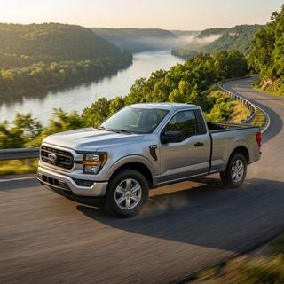 A cinematic, low-angle tracking shot of a silver 2026 Ford F-150 cruising confidently along a winding road atop the riverfront bluffs of the Ohio River valley, lush green deciduous trees and steep hillside terrain in the background, late afternoon golden hour lighting casting a warm glow over the scene, a close-up focus on the front wheel where high-performance ventilated brake rotors and pristine calipers are visible through the intricate alloy spokes, the truck maintaining a safe and controlled posture on the crisp asphalt, atmospheric mist rising from the river below, photorealistic style, sharp details, vibrant natural colors, a sense of rugged reliability and family safety, professional automotive photography.