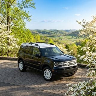 A photorealistic automotive lifestyle shot of the 2026 Ford Bronco Sport Heritage Edition, finished in black with a contrasting Oxford White roof and grille, parked on a scenic ridge overlooking the rolling green hills of the Ohio Valley in early May. The scene features the vibrant, fresh foliage of mid-spring, with lush maple trees and blooming white dogwood blossoms catching the soft morning sunlight. The composition is a dynamic three-quarter front angle, capturing the vehicle’s rugged 4WD stance and specialized suspension against a backdrop of the winding roads near Boardman and Calcutta. The sky is a brilliant clear blue with wispy white clouds, casting gentle reflections on the SUV’s polished surfaces. The atmosphere is crisp and bright, reflecting a perfect spring day for a family adventure, cinematic lighting, shallow depth of field, high-resolution photography, natural textures of the paved overlook and surrounding forest greenery.