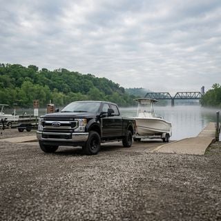 2026 Ford F-250 Super Duty XL, black paint, parked on a gravel landing beside a concrete boat ramp at an Ohio River marina in East Liverpool, professional 4WD truck, towing a white fiberglass fishing boat on a dual-axle trailer, 6.7L V8 diesel engine power, realistic working-class environment, spring morning in May, soft diffused sunlight through a thin layer of stratus clouds, lush green deciduous trees on the distant river bluffs, moist air, slight morning mist on the river surface, industrial steel bridge visible in the hazy background, wide-angle ground-level shot, sharp focus on the truck’s chrome-accented grille and heavy-duty tires, rugged and practical atmosphere, authentic tri-state regional vibe. ford and f-250 visible