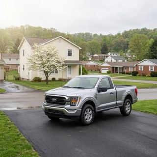 A 2026 Ford F-150 parked on a clean, paved driveway of a modest, well-maintained two-story frame house in a quiet Calcutta, Ohio suburban neighborhood, eye-level wide angle shot, soft May afternoon sunlight filtering through a light spring haze, the truck is the clear focal point with a polished finish reflecting the overcast sky, gently rolling hills and patches of fresh green woodland in the background, a small flowering dogwood tree in the front yard with white blossoms, a damp asphalt street and a neighborhood of mid-century suburban homes, cool spring air, naturalistic and crisp photography style, realistic and practical working-class setting, no text.
