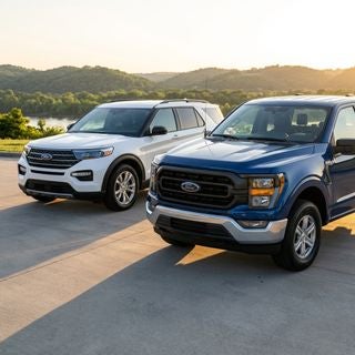Cinematic commercial photography, wide-angle ground-level shot, a pristine 2025 Ford F-150 in iconic deep blue and a white 2025 Ford Explorer, parked prominently on a polished dealership lot, set against the soft rolling hills and lush greenery of the Ohio River Valley, warm golden hour sunlight casting long soft shadows across the pavement, bright and inviting atmosphere, conveying transparency and community trust, hyper-realistic textures, vibrant natural colors, professional automotive lighting, high-gloss reflections on the vehicle bodies, shallow depth of field focusing on the front grills of the vehicles, crisp and clean aesthetic.