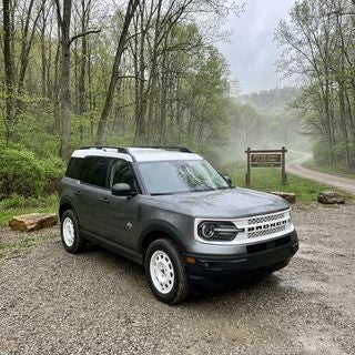 2026 Bronco Sport, adventure-ready compact SUV, parked on a damp, gravel trailhead parking lot near Beaver Creek, Calcutta, Ohio, square-shouldered design, rugged grey metallic paint, standard 4WD hardware visible, wide-angle ground-level shot, early May morning, soft overcast spring sky, patches of morning mist, surrounding forest of budding maple and oak trees, fresh green undergrowth, damp earth, wet sandstone rocks, realistic rural Ohio landscape, background features a simple wooden park sign and a winding dirt path into the rolling hills, cool ambient light, crisp reflections on the hood, quiet outdoor mood, practical suburban adventure style.