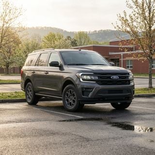 A 2026 Ford Expedition in a practical Carbonized Gray finish, parked on a clean asphalt school parking lot in Calcutta, Ohio, late afternoon in mid-May, blooming spring greenery and budding maples in the background, soft golden hour sunlight filtering through a light spring haze, the full-size SUV is the clear focal point, low-angle three-quarter view highlighting its massive presence and high ground clearance, realistic Appalachian Plateau terrain with gently rolling hills in the distance, modest suburban architecture of a public middle school with brick facades, damp pavement from a recent light spring shower, reflective puddles on the ground, muted but warm color palette, sharp focus on the vehicle’s modern headlights and front grille, high-detail automotive photography style.