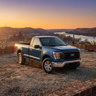 Cinematic, photorealistic wide shot of a modern Ford F-150 pickup truck, low-angle perspective emphasizing rugged durability and power, parked on a dusty construction job site overlooking the rolling hills and weathered industrial landscape of the Ohio River valley, golden hour sunlight casting a warm amber glow across the metallic body, long shadows, subtle lens flare, crisp textures of gravel and weathered brick in the background, distant Appalachian architecture and steel bridge silhouettes, a mood of hardworking dependability and pride in American craftsmanship, hyper-detailed automotive photography, rich color palette of deep metallic blue and chrome accents against a vibrant orange and purple sky, high-resolution, professional commercial lighting.