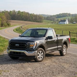 Ford F-150, current model, iconic American pickup truck, parked on a gravel shoulder of a two-lane road in rural Beaver Township, Ohio, May morning light, soft spring sunshine filtering through a light haze, gently rolling hills of the Appalachian Plateau in the background, fertile agricultural fields with fresh green sprouts, patches of budding hardwood forest, a classic wooden fence line, mid-ground features a small white local church with a modest steeple, the truck is clean but clearly a tool for work, metallic paint reflecting the pale blue sky and soft clouds, low-angle hero shot, focus on the rugged front grille and headlamps, color palette of earthy greens, soft browns, and deep blues, peaceful and dependable mood, realistic photography style, reflecting a quiet and hardworking Midwestern spring morning.