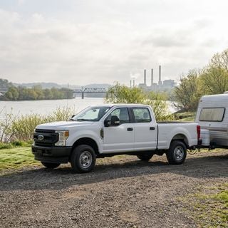 2026 Ford F-250 Super Duty, Oxford White XL trim, 6.7L V8 Diesel, parked on a gravel landing at a practical Ohio River camping spot in East Liverpool, wide-angle shot, side profile highlighting the massive towing hitch and 4WD stance, May morning light, soft spring sunshine, thin hazy clouds, budding willow trees and fresh green brush along the riverbank, steel-grey water of the Ohio River in the background, a modest camper on a trailer hitched to the rear, industrial bridge and distant manufacturing stacks on the horizon, realistic working-class atmosphere, calm and productive mood, photorealistic, sharp textures on the chrome grille and rugged tires, morning dew on the grass, no people, no text.