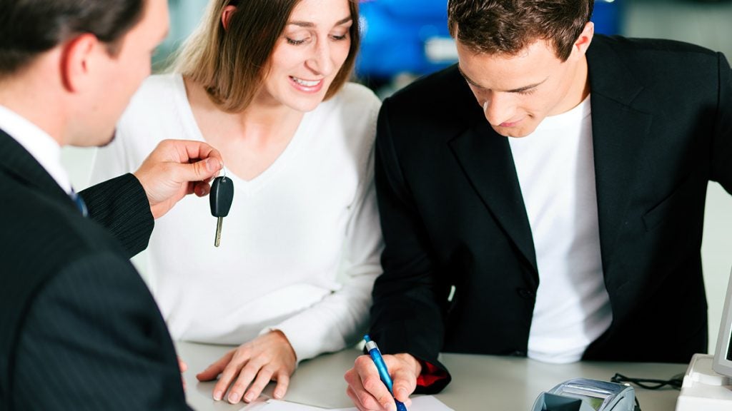 A car salesperson hands keys to a couple as the man signs paperwork at a dealership desk.