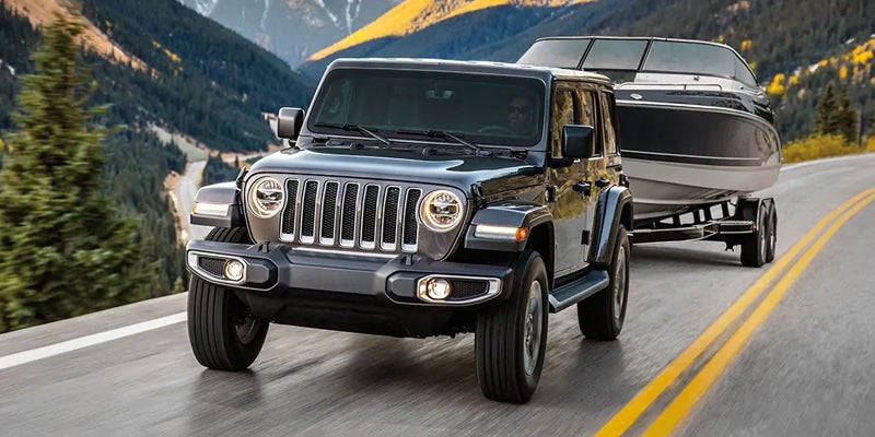 A jeep tows a boat on a trailer up a winding paved road with mountains and forests in the background.