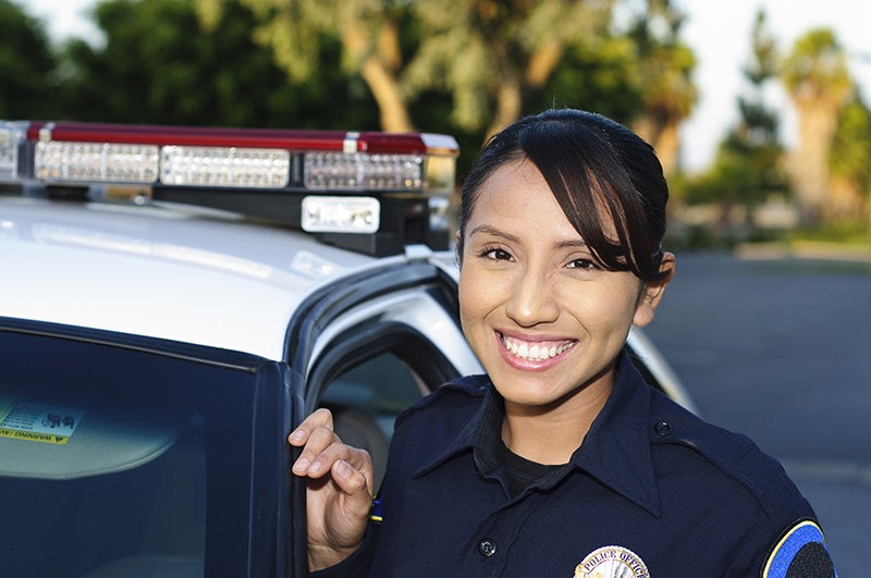 A smiling female police officer stands next to a patrol car with flashing lights.