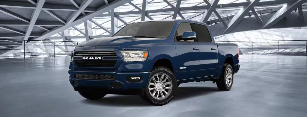 A dark blue Ram 1500 truck with chrome wheels is parked on a glossy floor in a modern showroom with large windows and a gray truss ceiling.