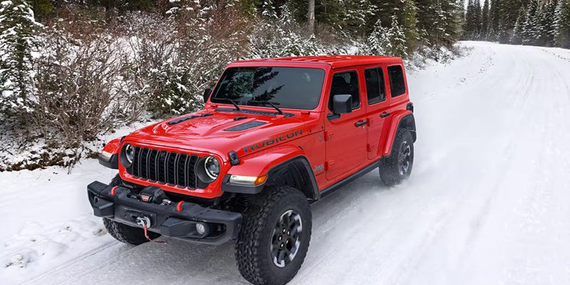 A red Jeep Wrangler Rubicon drives on a snowy road through a forest.