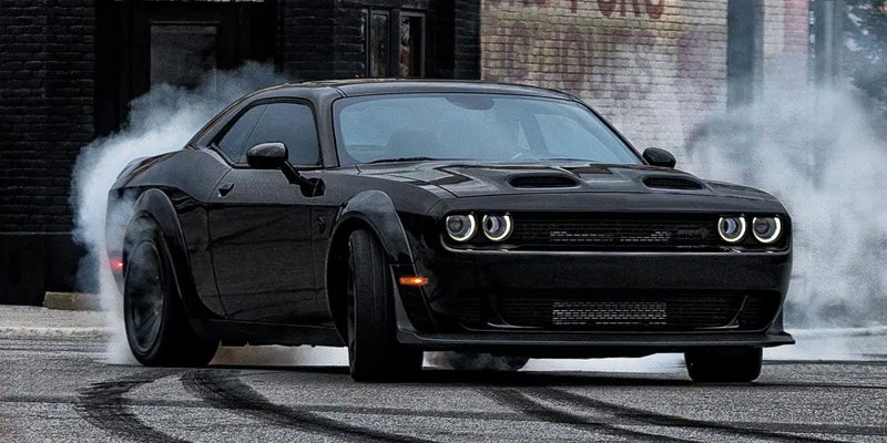 Black Dodge Challenger Hellcat making a burnout, smoke rising from tires on asphalt, red brake lights.