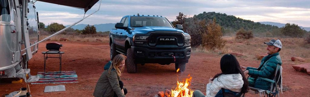 People around a campfire with a Ram truck, a silver camper, and a grill in a rustic outdoor setting.