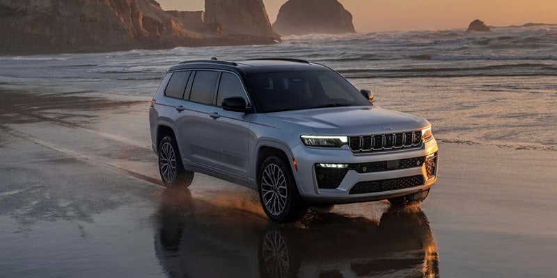 Silver Jeep Grand Cherokee L driving on a wet sandy beach with ocean waves in the background.