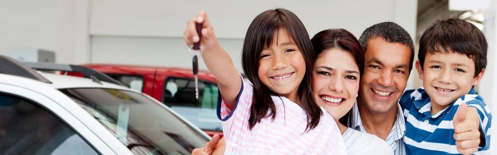 A smiling family—father, mother, daughter, and son—stands in front of cars. The daughter holds up a car key.