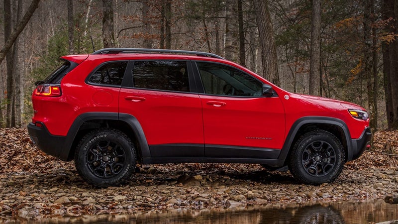 Left side of a red Jeep Cherokee pulling a trailer through a forest with leaves on the ground.