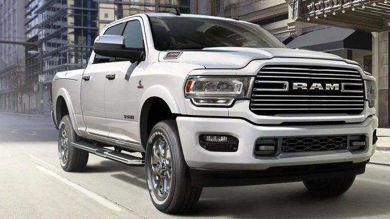 A white Ram 2500 pickup truck drives on a city street. Tall buildings are in the background.