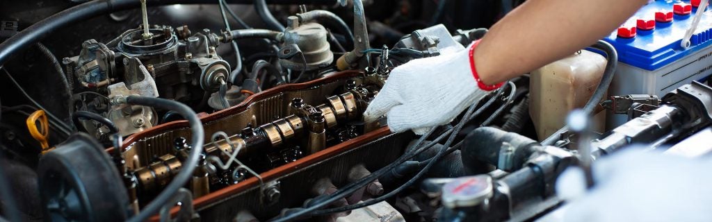 A gloved hand works on an open car engine, revealing the camshaft and other internal components. A battery is visible.