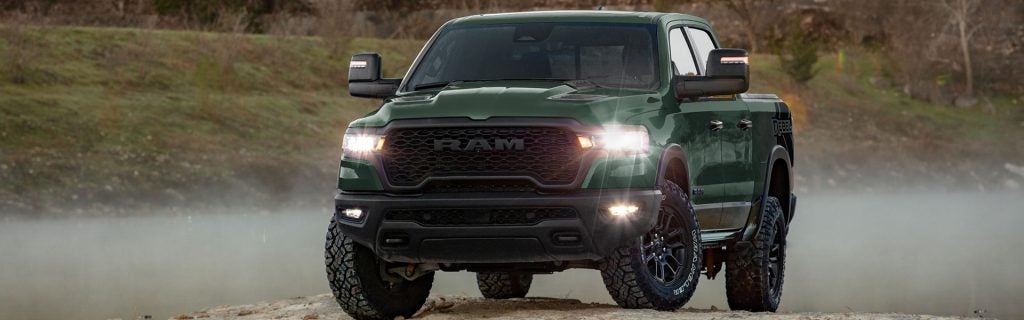A dark green Ram truck facing forward, with its headlights brightly lit, on a dirt path with fog rolling in.
