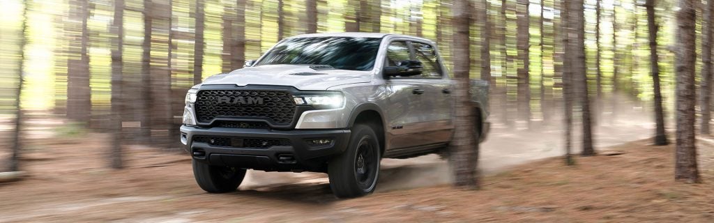 A gray Ram truck drives quickly on a dirt road through a forest. Trees are blurred due to speed.