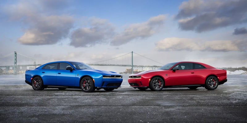 A blue four-door car and a red two-door car parked side-by-side on asphalt, with a suspension bridge in the background.