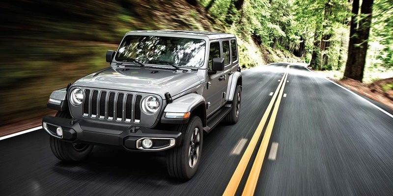 A gray Jeep Wrangler driving fast on a winding road through a forest. Trees are blurred due to speed.