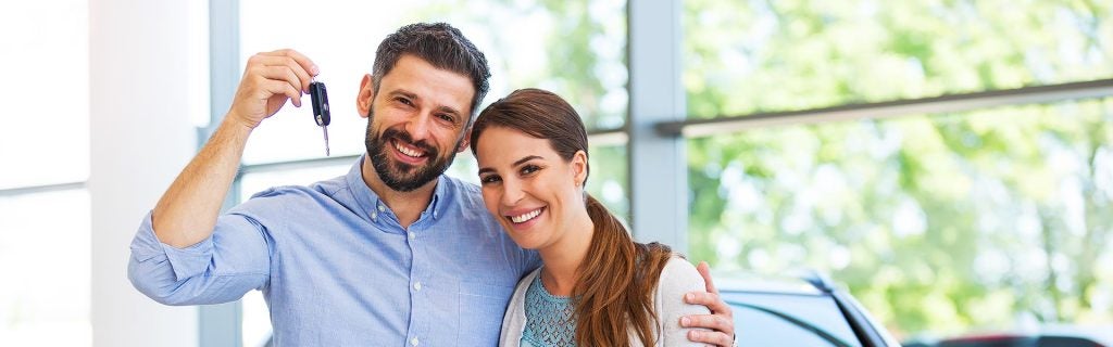 A couple, smiling, holding a car key, in a dealership. The man holds the key, the woman leans into him.