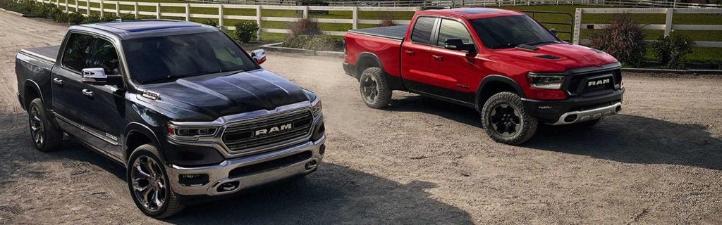 Two Ram pickup trucks, one black and one red, parked on a dirt path with a white fence in the background.