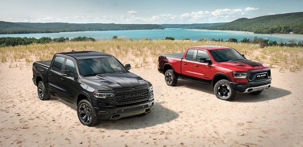 Two Ram pickup trucks, one black and one red, parked on a sandy dune. A lake and forested hills are in the background.