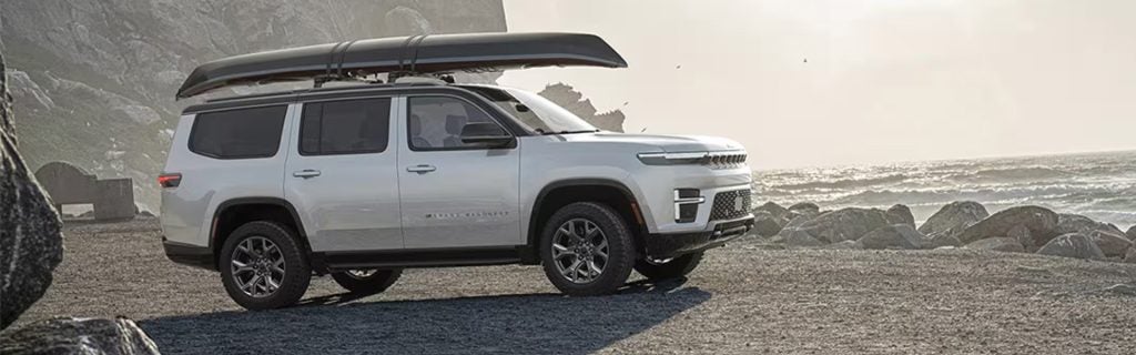 Silver SUV with a canoe on its roof rack, parked on a rocky beach next to the ocean.